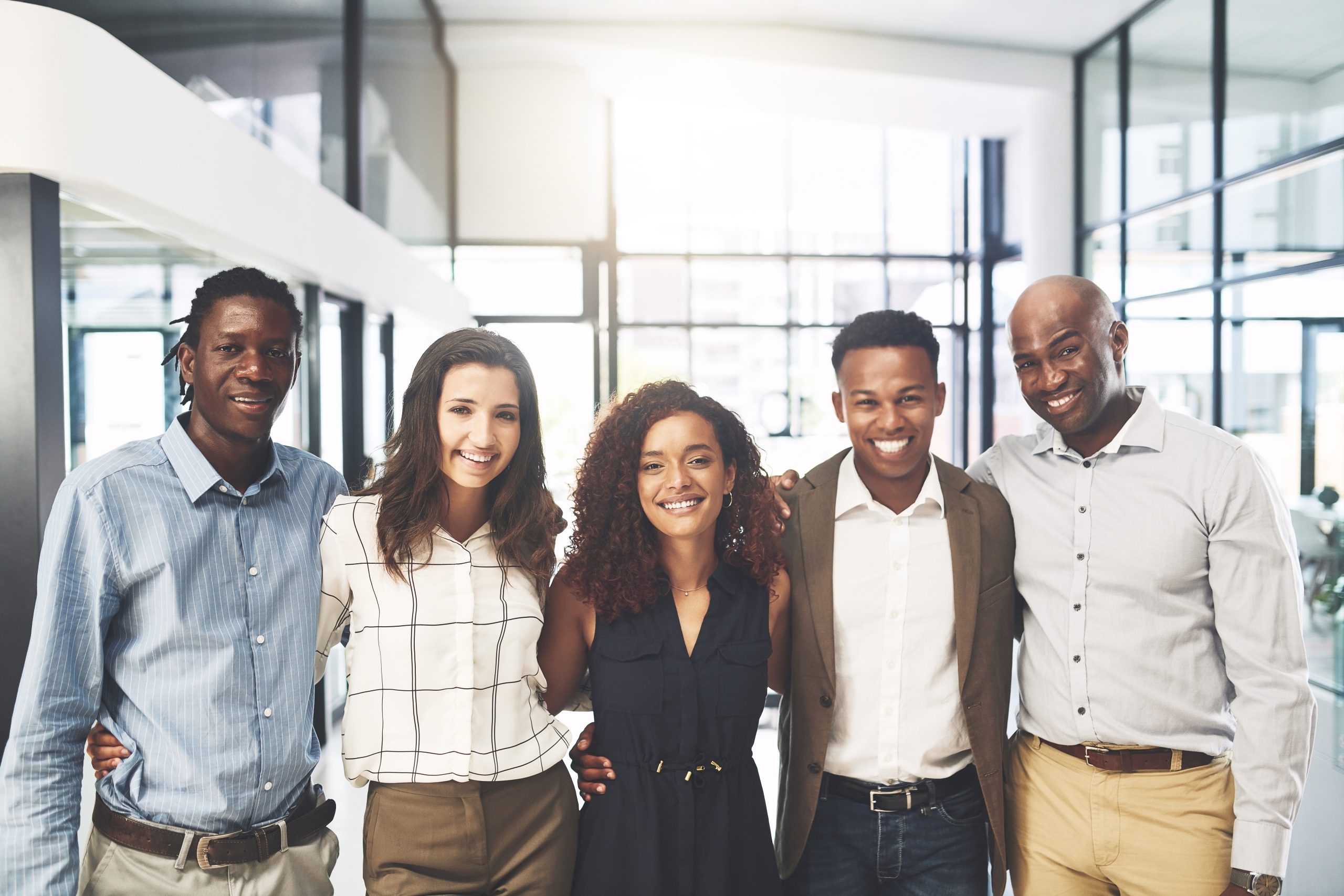 Five people stand close together in an office hallway, smiling at the camera. They appear friendly and happy, with arms around each other, suggesting teamwork and camaraderie in a professional environment.