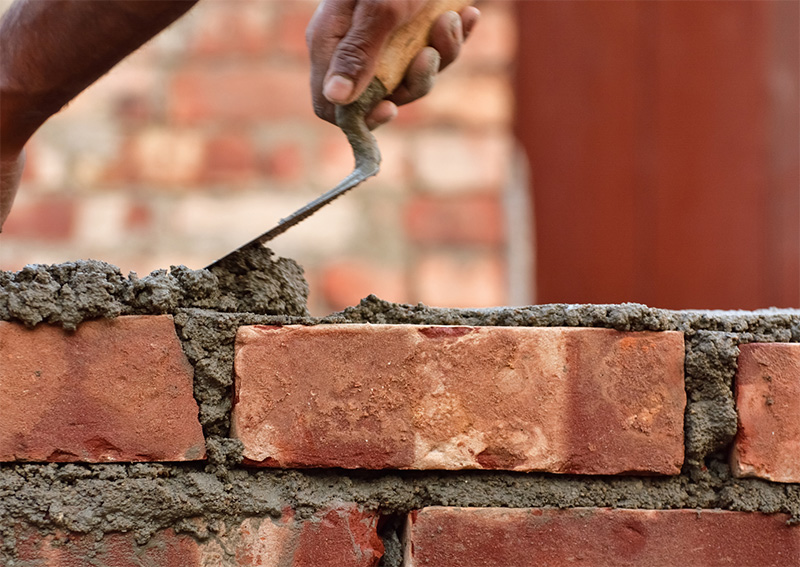 A close-up of a persons hand using a trowel to spread mortar between red bricks while constructing a brick wall.