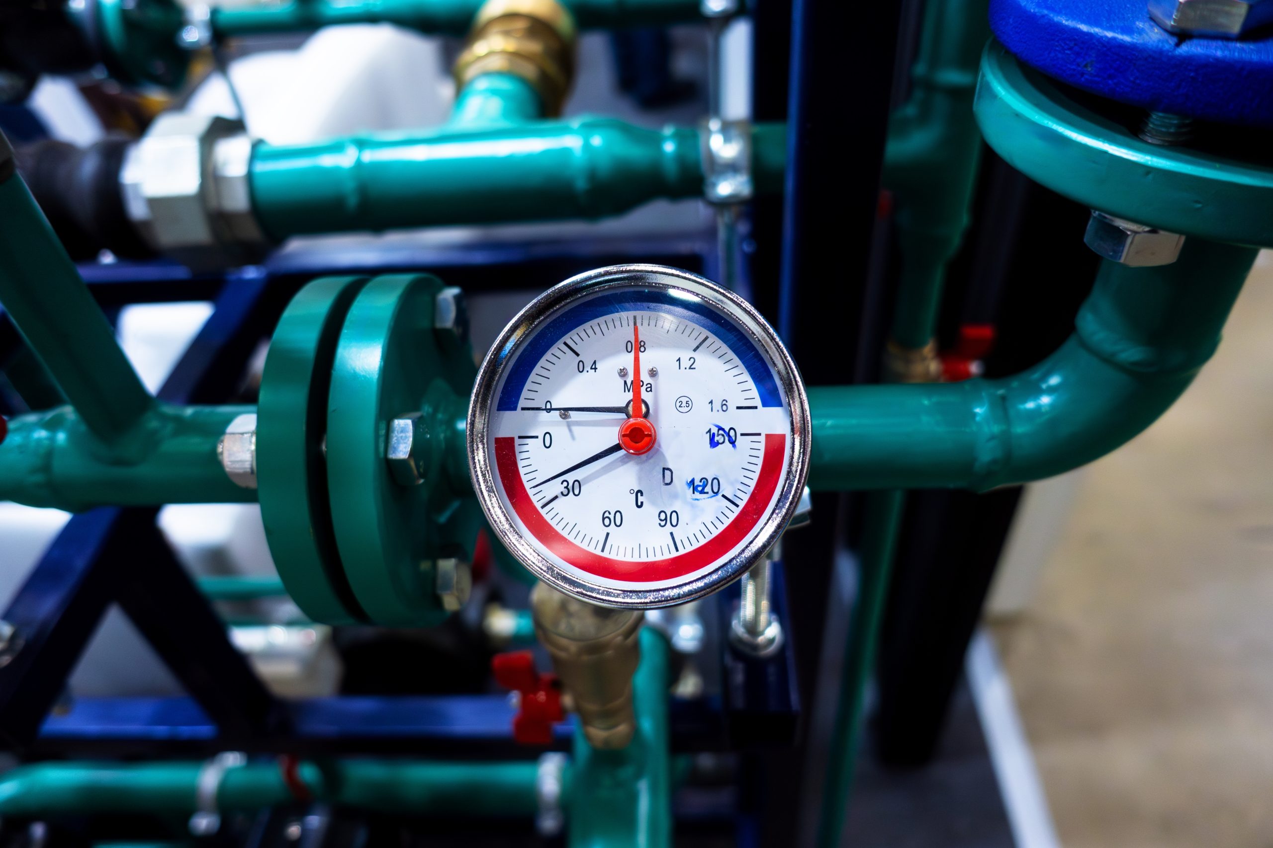 A close-up of a round pressure gauge attached to green metal pipes, displaying a reading of approximately 2.7 bar, with both blue and red sections indicating pressure levels.